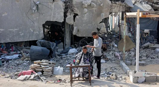 Un uomo taglia i capelli a un altro davanti a un edificio distrutto a Jabalia, nel Nord della Striscia di Gaza